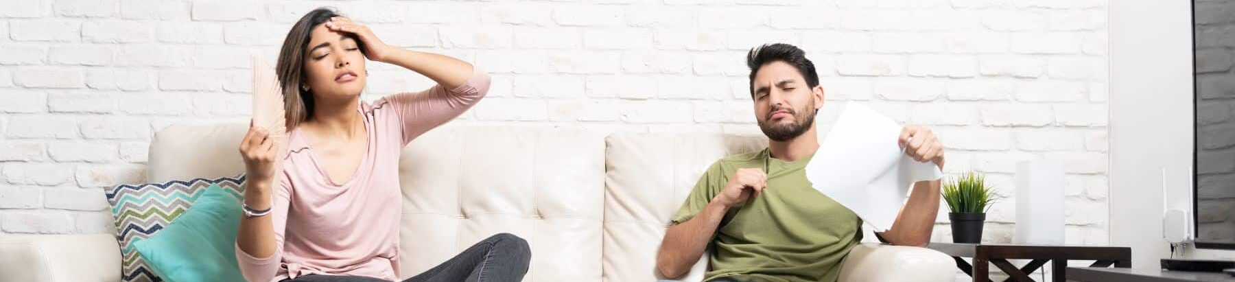 Man and woman sitting on a couch in a living room, both looking overheated. The woman is fanning herself with a piece of paper while the man is pulling at his shirt collar and also holding a piece of paper, indicating discomfort from high temperatures