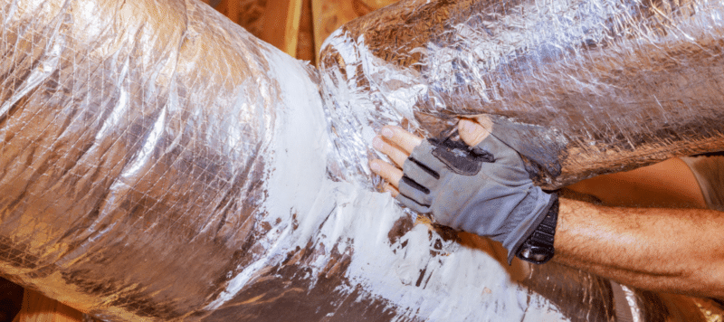 A technician wearing work gloves applies sealant to insulated ductwork during a repair or replacement process